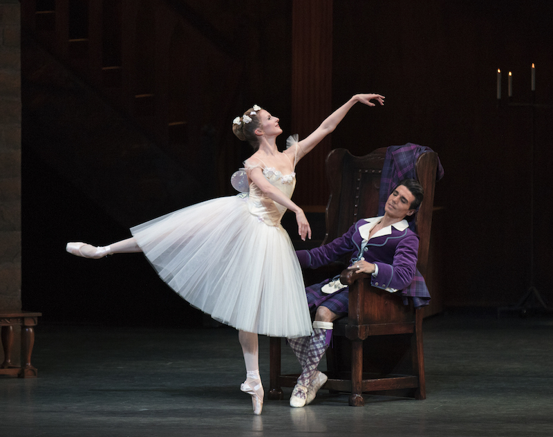 Sterling Hyltin performs a Romantic style arabesque on point while Joaquin De Luz dressed in a bright purple kilt lazily sits on a throne